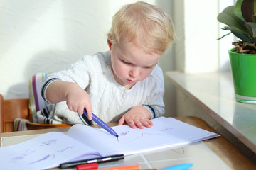 Naklejka premium blonde toddler girl drawing family while sitting indoors