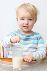 Cute blonde toddler girl drinking milk from glass with straw