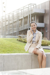 Happy young businesswoman using mobile phone while sitting on wall against office building