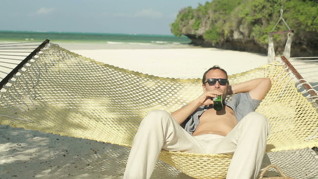 Happy man on hammock raising toast to camera with bottle of beer