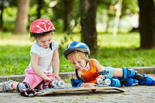 Little boy and girl in roller equipment sit on walkway in summer