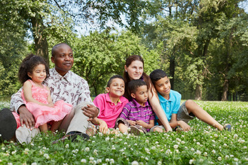 Happy interracial family of six sits on grass on lawn in park