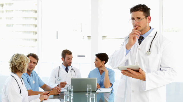 Thoughtful doctor using tablet with staff talking behind him
