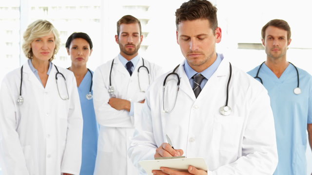 Serious doctor writing on clipboard while staff standing behind