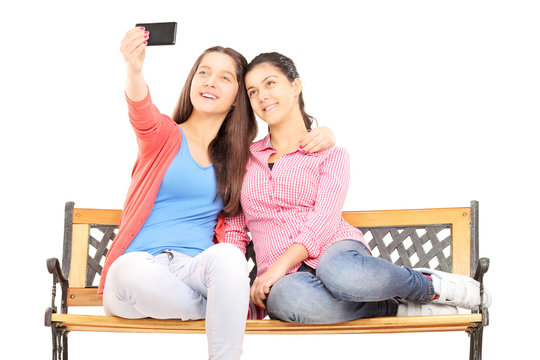 Two young girls seated on bench taking picture of themselves wit