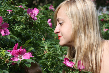 Beautiful blonde girl looks at pink flowers on bush in park