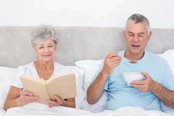 Senior couple with book and cereal bowl in bed