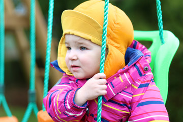 toddler girl in multicolor jacket and orange hat rocks on swing