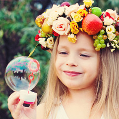 little girl with wreath from flowers blows soap bubbles
