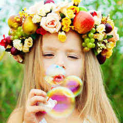 little girl with wreath from flowers blows soap bubbles