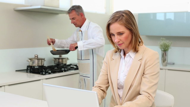 Businesswoman using laptop while husband cooks dinner