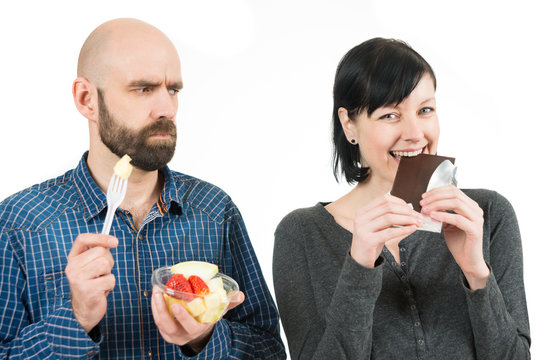 Mann mit Obst schaut b&ouml;se zu Schokolade essender Frau