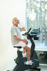 Tired senior man with towel on exercise bike in fitness club