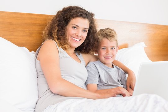 Smiling mother and son sitting on bed with laptop