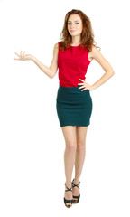 Obraz premium Confident young woman in red top and green skirt presenting product or service, full body isolated on white background, she has curly brown hair and is wearing black high heels