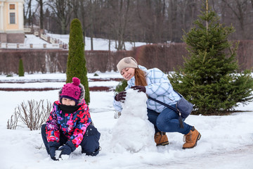 Young mother and daughter making snowman with snow in winter park