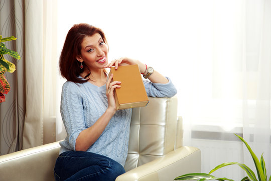 Cheerful woman sitting on the sofa and hlding a book at home