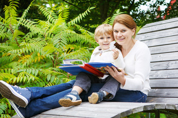 Little boy and his mother sitting on bench in park and reading b