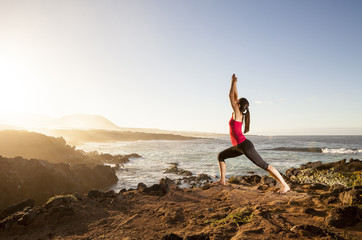 Fototapeta na wymiar Young woman practicing yoga warrior pose near the ocean at sunse