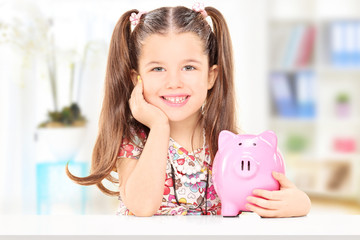 Cute girl sitting at table and holding a piggybank at home