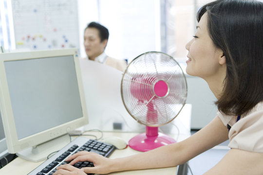 Woman Using PC On Desk With An Electric Fan