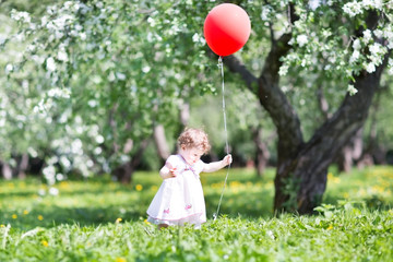 Funny baby girl walking in an apple tree garden with a red ballo