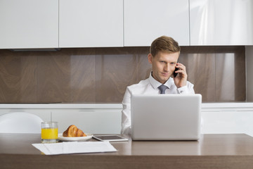 Mid adult businessman on call while using laptop at breakfast table