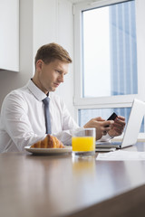 Mid adult businessman using cell phone with laptop on breakfast table
