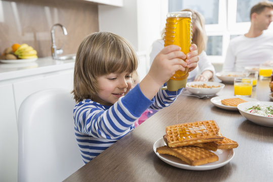 Happy Boy Pouring Honey On Waffles While Having Breakfast With Family