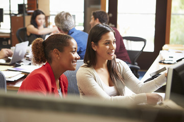 Two Colleagues Working At Desk With Meeting In Background