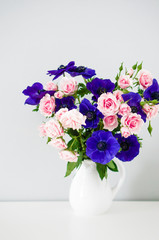 Bouquet of pink roses and blue anemones in white vase on grey ba