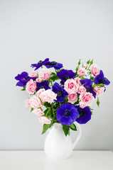 Bouquet of pink roses and blue anemones in white vase on grey ba