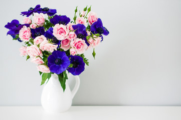 Bouquet of pink roses and blue anemones in white vase on grey ba
