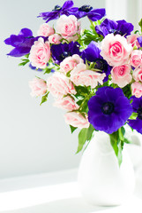 Bouquet of pink roses and blue anemones in white vase near the w