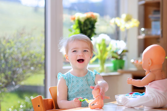  toddler girl feeding with yogurt her dolls sitting in kitchen