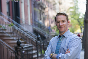 A Man In A Blue Shirt And Blue Tie With Arms Folded, Standing On The Sidewalk