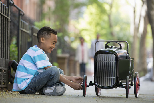 A Young Boy Playing With A Old Fashioned Toy Car On Wheels On A City Street. 
