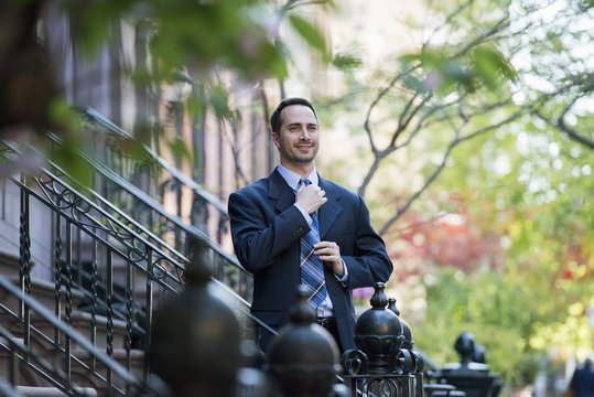 A Man In A Business Suit Adjusting His Tie. At The Bottom Of The Steps Of A Townhouse In A Terrace.