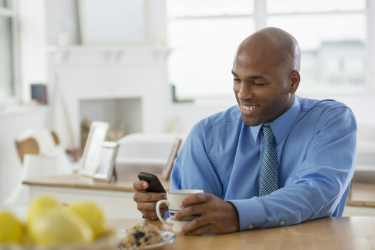 A Man In A Blue Shirt, Sitting At A Breakfast Bar Using A Smart Phone.
