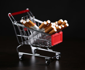 Cigarettes in shopping cart on wooden table on dark background