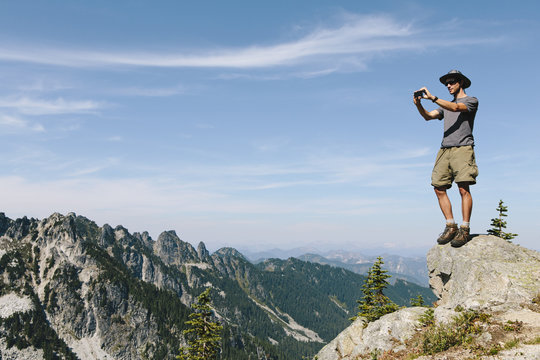 A hiker on a mountain summit, holding a smart phone, at the top of Surprise Mountain, in the Alpine Lakes Wilderness, in Mount Baker-Snoqualmie National Forest.