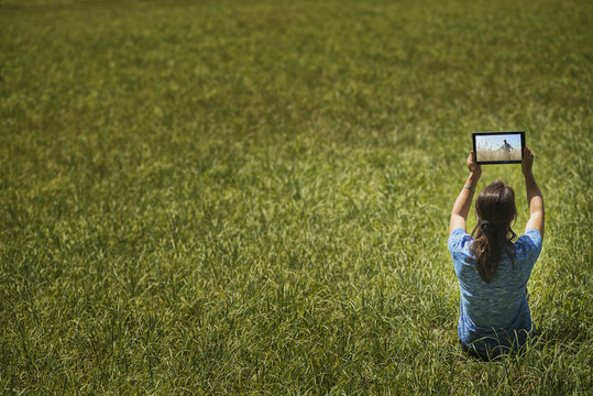 View from above of a woman sitting on grass, lifting up a personal computer notepad, looking at an image. 