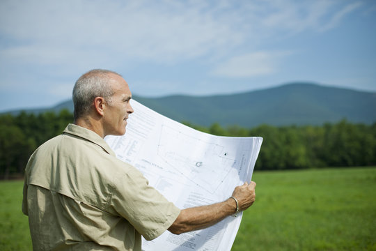 A rural scene and mountain range, and a person holding a scale model of a new building.