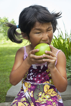 A small child with pigtails chewing a large green apple.