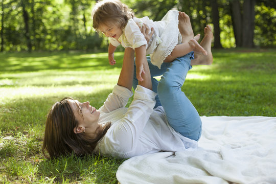 A mother lying on her back with her twin daughter balancing on her knees, outdoors. 