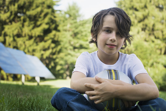 A boy holding a football, sitting on the grass. Solar panels. 