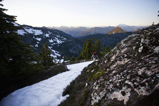 A young man scrambles around a snow patch while hiking to the summit of a large mountain in the Cascades of Washington, USA.
