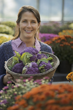 A woman holding a bowl of fresh produce, purple sprouting broccoli. Flowering plants. Crysanthemums.