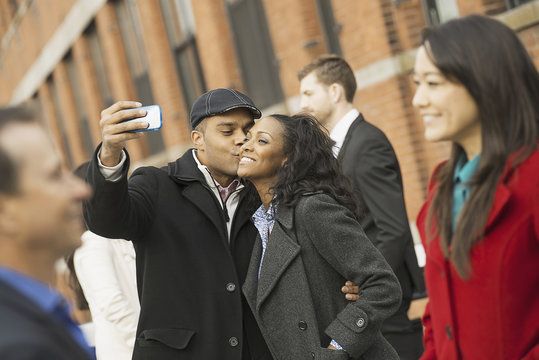 City life. A group of people on the go. A man holding out a camera phone and taking pictures of the group. Kissing a young woman. Men and women.
