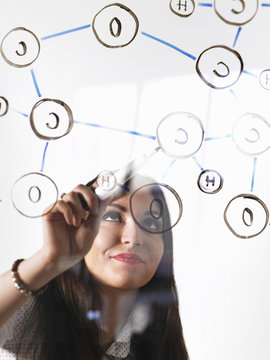 A young woman writing a chemical notation for the gas Oxygen, the molecular structure, on a clear seethrough panel with a black pen.